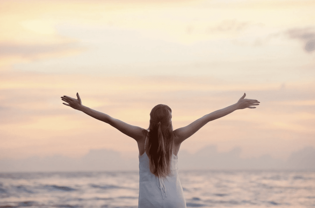 Woman holding her hands up in the air in a victory pose with an ocean horizon in front of her.